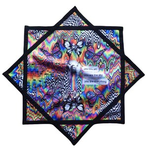 Puede incluir: Una colorida pieza de arte textil en forma de estrella con un diseño psicodélico. Presenta mariposas, una figura central y el texto "you are art", "you are the sky" y "you are everything". El borde es negro.