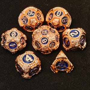 May include: Set of seven copper and blue metal dice with intricate gear designs and gold numbers. The dice are a d4, d6, d8, d10, d%, d12, and d20.