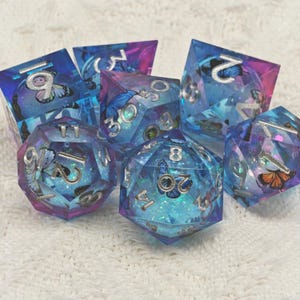 May include: A set of seven iridescent blue and purple dice with silver numbers and butterfly designs. The dice are made of clear resin with glitter embedded in the material.