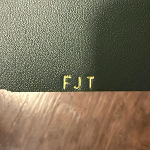 May include: Close-up of a dark green leather surface with the gold letters "FJT" embossed on the top. The leather has a textured appearance. The bottom of the image shows a wooden surface.