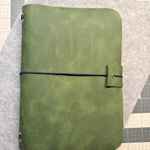 May include: A green leather journal with a textured surface and a dark blue elastic closure. The journal is closed with a matching tab and secured with a dark blue elastic band. The journal is lying on a gray felt surface.