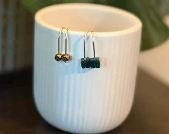 May include: Two pairs of U-shaped drop earrings. One pair features small, metallic gold-toned geometric cubes, while the other pair showcases deep emerald green translucent square beads. The earrings are displayed on the rim of a white ribbed cup.