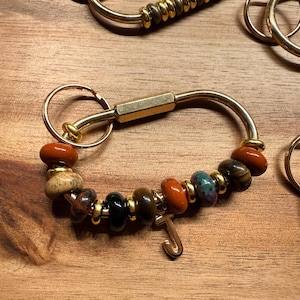 May include: A gold-toned keychain with a curved bar and a ring. The keychain is adorned with a series of colorful beads in shades of brown, orange, and green. A gold-toned letter "J" charm hangs from the beads.
