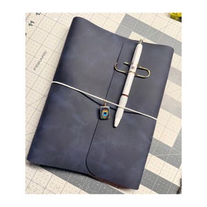 May include: A dark blue leather journal with a white elastic band and a white pen secured by a gold-colored pen holder. A small, colorful pendant hangs from the elastic. The journal is closed and appears to be new.