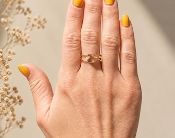May include: A gold-toned wire ring featuring a central, small, speckled beige stone bead. The ring is constructed from thin, coiled metal wire that forms an open, oval-shaped frame around the bead, creating a delicate and minimalist jewelry piece.