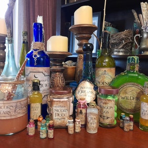 May include: A collection of glass bottles with various labels, including "Unicorn Tears", "Beetle Juice", "Unicorn Poo", "Bottled Dreams", "Dragon Eggs", "Wildfire", and "Moon Dust". The bottles are arranged on a wooden surface with candles and other decorative items.
