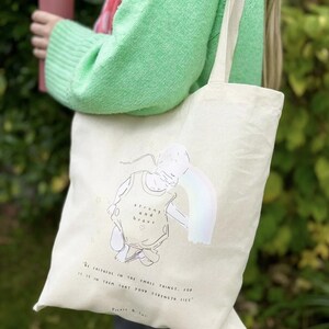 Puede incluir: Bolso tote de color crema con un dibujo lineal de una persona y un arcoíris. La bolsa tiene las palabras "fuerte y valiente" y la cita "Sé fiel en las pequeñas cosas, porque en ellas reside tu fuerza."