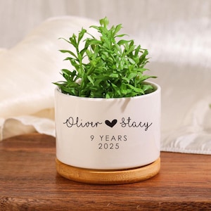 Op de afbeelding: Witte keramische plantenpot met een bamboe basis. De pot heeft zwarte tekst met de tekst "Oliver ♥ Stacy 9 YEARS 2025".