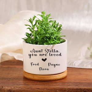May include: A white ceramic planter with a bamboo base, holding a green plant. The planter has the text "Aunt Stella, you are loved" with names below. The planter sits on a wooden cutting board.