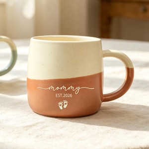 May include: A two-toned ceramic mug with a cream-colored top and a terracotta-colored bottom. The front features the white text "mommy", "EST.2026", and a small graphic of two baby footprints. A second, partially visible mug is in the background.