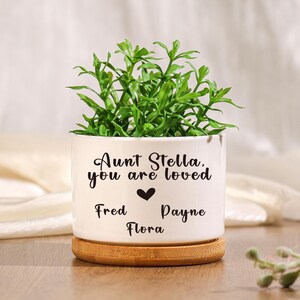 Può includere: Un vaso di ceramica bianco con la scritta "Aunt Stella, you are loved" e nomi sottostanti. Il vaso contiene una pianta verde e poggia su una piccola base di legno. Un simbolo a forma di cuore è tra il testo e i nomi.