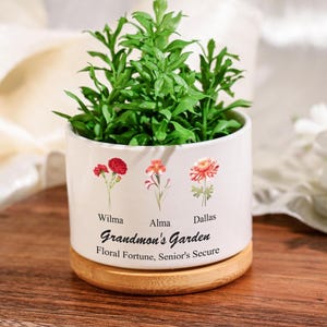 Puede incluir: Maceta de cerámica blanca con base de madera. La maceta tiene tres flores con los nombres Wilma, Alma y Dallas impresos debajo. El texto "Grandmon's Garden" está impreso debajo de las flores, y el texto "Floral Fortune, Senior's Secure" está impreso debajo de ese.
