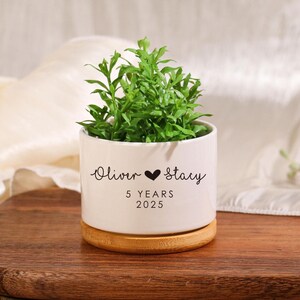 May include: White ceramic planter with a bamboo base. The planter has a black inscription that reads "Oliver ♥ Stacy 5 YEARS 2025". A small green plant is growing in the planter.
