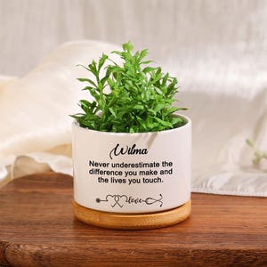 May include: White ceramic planter with a bamboo base. The planter has a green plant inside and the text "Wilma Never underestimate the difference you make and the lives you touch." printed on the front.