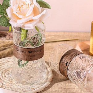 Könnte beinhalten: Eine klare Glasvase mit strukturiertem Design enthält drei pfirsichfarbene Rosen mit grünen Blättern. Ein braunes Band mit Text umschließt die Vase. Eine zweite Vase liegt auf der Seite. Ein gewebter Untersetzer und ein Band befinden sich im Vordergrund.