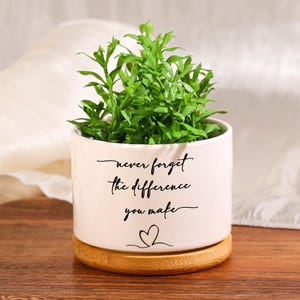 Puede incluir: Una maceta de cerámica blanca con el texto "never forget the difference you make" y un símbolo de corazón, que contiene una planta verde. La maceta se encuentra sobre una base de madera y una bandeja de madera.