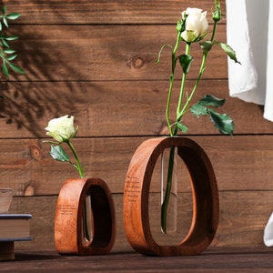 May include: Two wooden vases with glass tubes for holding flowers. The vases are oval shaped and have a natural wood finish. One vase holds a single white rose, while the other holds a bouquet of white roses.