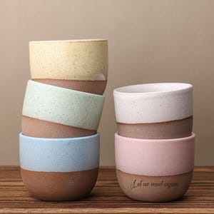 May include: Six ceramic cups in pastel shades, including yellow, mint, light blue, pink, and white. Each cup has a speckled upper section and a brown base. The bottom cup features the text "Let us meet again".