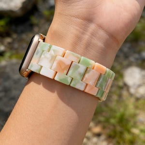 May include: A gold Apple Watch with a colorful watch band. The band is made of rectangular links in shades of white, green, and peach. The watch is on a person's wrist.