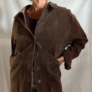May include: A brown suede jacket with a button-up front and a collared neckline. The jacket has long sleeves and two front pockets. The model is wearing a brown top and light-colored pants. The jacket appears to be vintage.