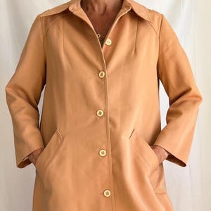 May include: A tan-colored trench coat with a collared neckline and button-down closure. The coat features two front pockets and is worn over blue jeans. The coat is a neutral color and has a classic design.