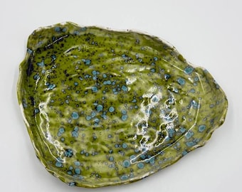 May include: A handmade ceramic oyster-shaped dish. The dish features a speckled green glaze with blue accents and a white rim. The organic shape and unique glaze make it a decorative piece for the home.