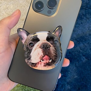 以下が含まれることがあります： フレンチブルドッグの電話グリップが付いた黒いスマートフォン。 電話グリップは、舌を出した黒、白、茶色のフレンチブルドッグのクローズアップ画像が特徴です。 電話は手で持たれています。