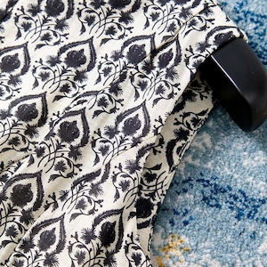 Op de afbeelding: Close-up van een cr&egrave;mekleurige stof met een herhalend zwart bloemenpatroon. Het ontwerp bevat gestileerde bladeren en bloemelementen. De stof is over een zwarte hanger gedrapeerd, wat op kleding duidt. De achtergrond is een blauw patroonkleed.