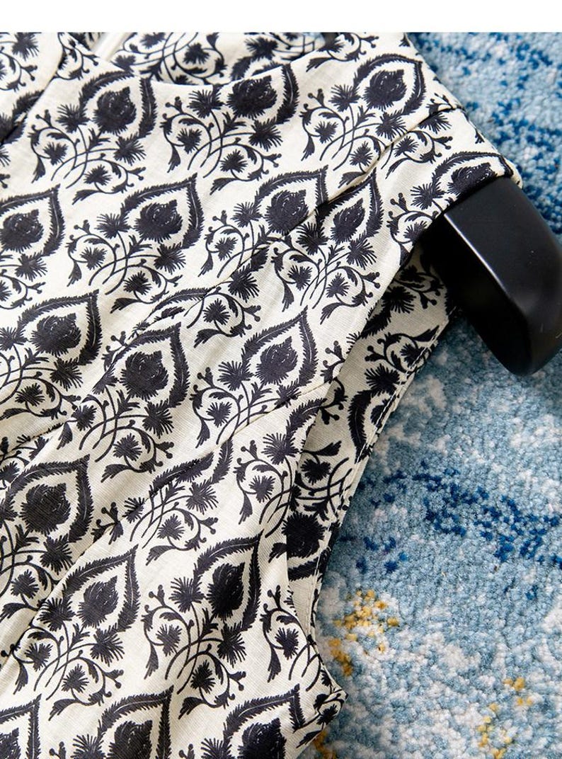 Op de afbeelding: Close-up van een cr&egrave;mekleurige stof met een herhalend zwart bloemenpatroon. Het ontwerp bevat gestileerde bladeren en bloemelementen. De stof is over een zwarte hanger gedrapeerd, wat op kleding duidt. De achtergrond is een blauw patroonkleed.