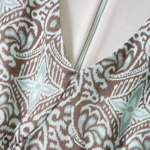 Op de afbeelding: Close-up van een V-hals kledingstuk met een bruin en lichtblauw paisley patroon. De stof heeft een gedetailleerd bloemmotief. Een zilveren rits loopt langs het midden van de halslijn, met een witte voering zichtbaar.