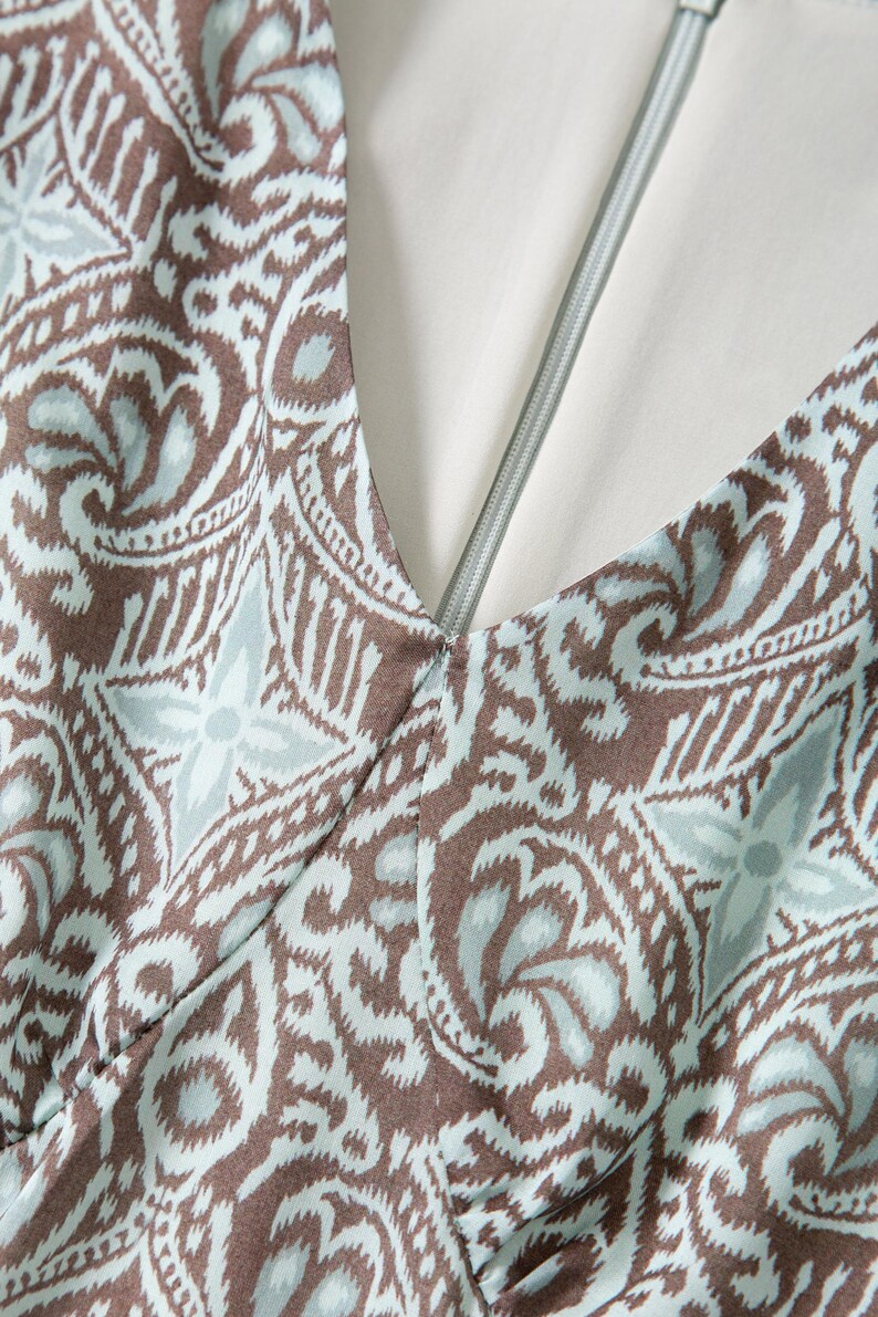 Op de afbeelding: Close-up van een V-hals kledingstuk met een bruin en lichtblauw paisley patroon. De stof heeft een gedetailleerd bloemmotief. Een zilveren rits loopt langs het midden van de halslijn, met een witte voering zichtbaar.