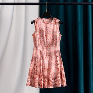 Op de afbeelding: Mouwloze jurk met een rood en wit stippenpatroon. De jurk hangt aan een zwarte hanger tegen een achtergrond van witte en teal gordijnen. De jurk heeft een getailleerd lijfje en een uitlopende rok.