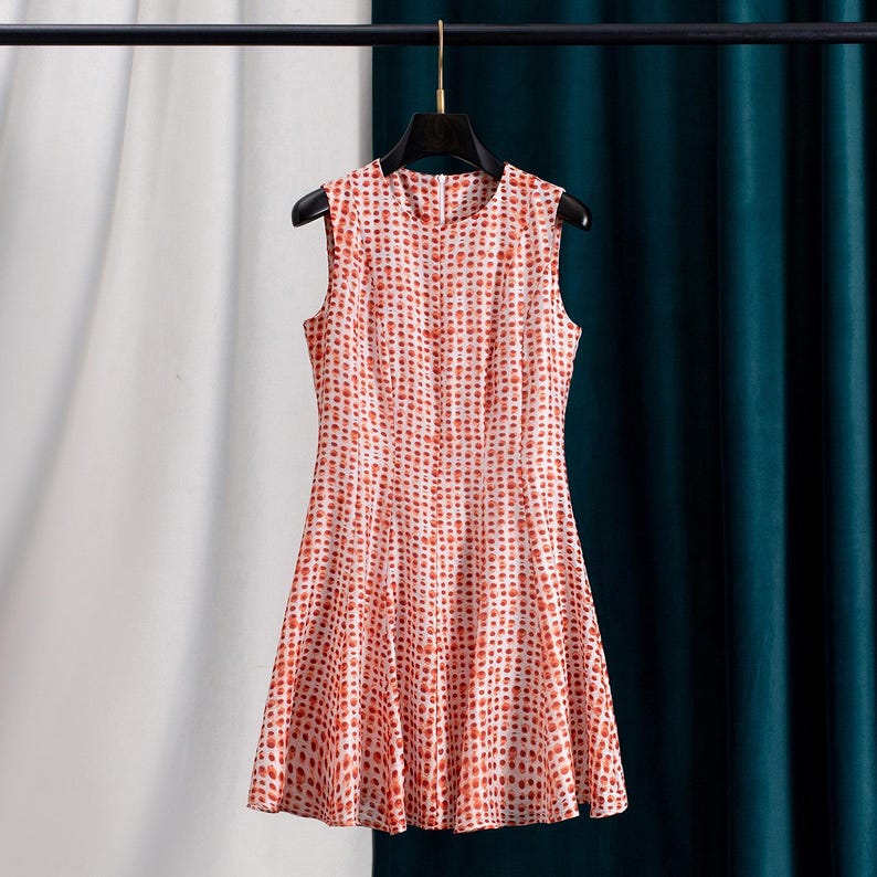 Op de afbeelding: Mouwloze jurk met een rood en wit stippenpatroon. De jurk hangt aan een zwarte hanger tegen een achtergrond van witte en teal gordijnen. De jurk heeft een getailleerd lijfje en een uitlopende rok.