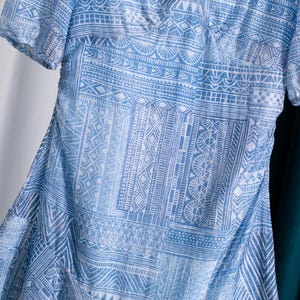 May include: A light blue dress with a white geometric pattern. The dress has short sleeves and a V-neck. The fabric appears to be lightweight and flowing, with a subtle sheen. The dress is partially visible, with a white and teal background.
