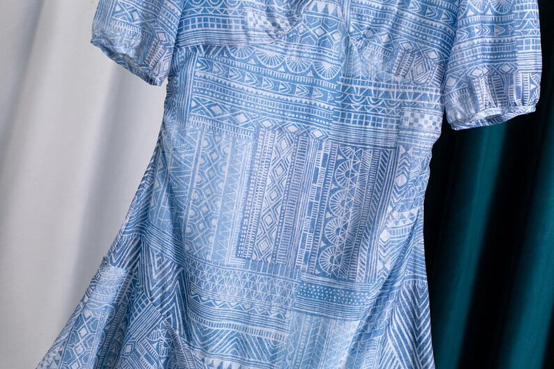 May include: A light blue dress with a white geometric pattern. The dress has short sleeves and a V-neck. The fabric appears to be lightweight and flowing, with a subtle sheen. The dress is partially visible, with a white and teal background.