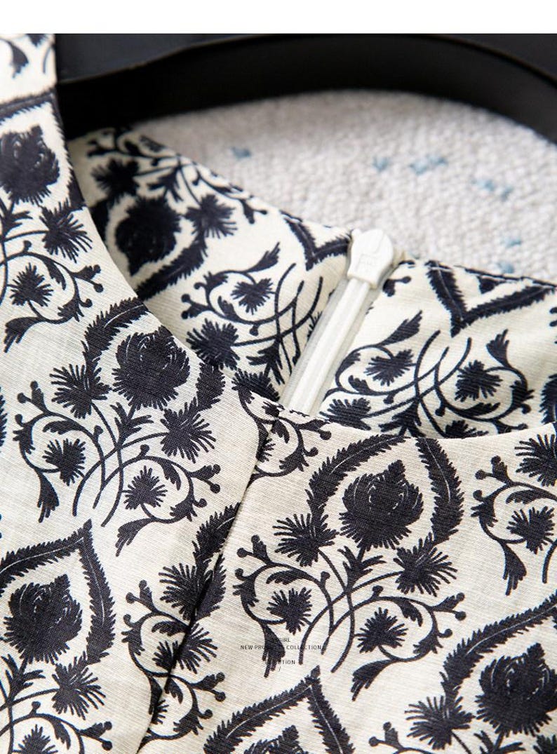 Op de afbeelding: Close-up van een cr&egrave;mekleurige stof met een zwart bloemenpatroon. Een witte rits is zichtbaar, wat suggereert dat het deel uitmaakt van een kledingstuk. Het ontwerp bevat gestileerde bloemen en bladeren.