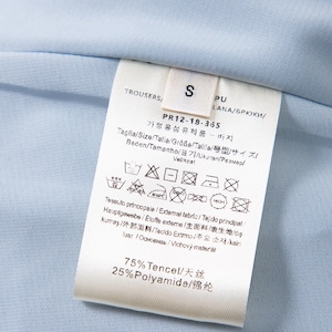 May include: Close-up of a light blue garment label with text and care symbols. The label indicates the garment is trousers, size S, and lists fabric composition as 75% Tencel and 25% Polyamide.