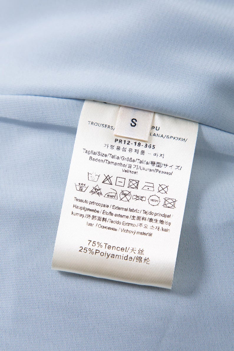 May include: Close-up of a light blue garment label with text and care symbols. The label indicates the garment is trousers, size S, and lists fabric composition as 75% Tencel and 25% Polyamide.