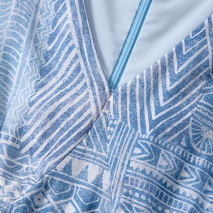 May include: Close-up of a light blue garment with a geometric white pattern. The fabric has a V-neck design with a light blue zipper. The overall aesthetic is a blend of modern and tribal influences, suitable for apparel.