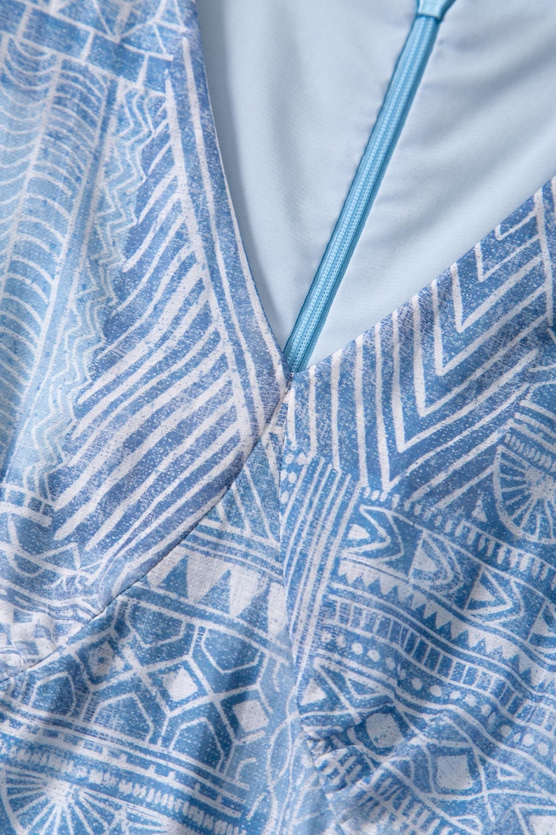 May include: Close-up of a light blue garment with a geometric white pattern. The fabric has a V-neck design with a light blue zipper. The overall aesthetic is a blend of modern and tribal influences, suitable for apparel.