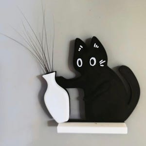 May include: A black cat-shaped shelf decor with a white vase holding tall, thin, gray-brown stems. The cat has white eyes, whiskers, and ear accents. The vase and shelf are white, and the background is a light gray.