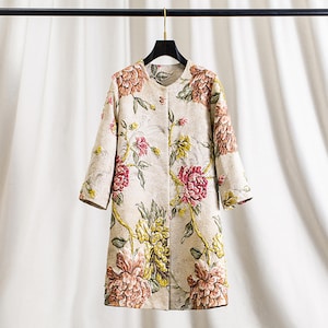 以下が含まれることがあります： クリーム色の花柄コート。丸襟と七分袖が特徴。ピンク、黄色、茶色の大きな花と緑の葉が繰り返されるパターン。黒いハンガーにかかっています。エレガントな衣服です。