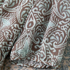 Op de afbeelding: Close-up van stof met een herhalend paisley-patroon in bruin en lichtblauw. De stof is geplooid en toont de mouw van een kledingstuk. Het patroon bevat bloem- en geometrische vormen.