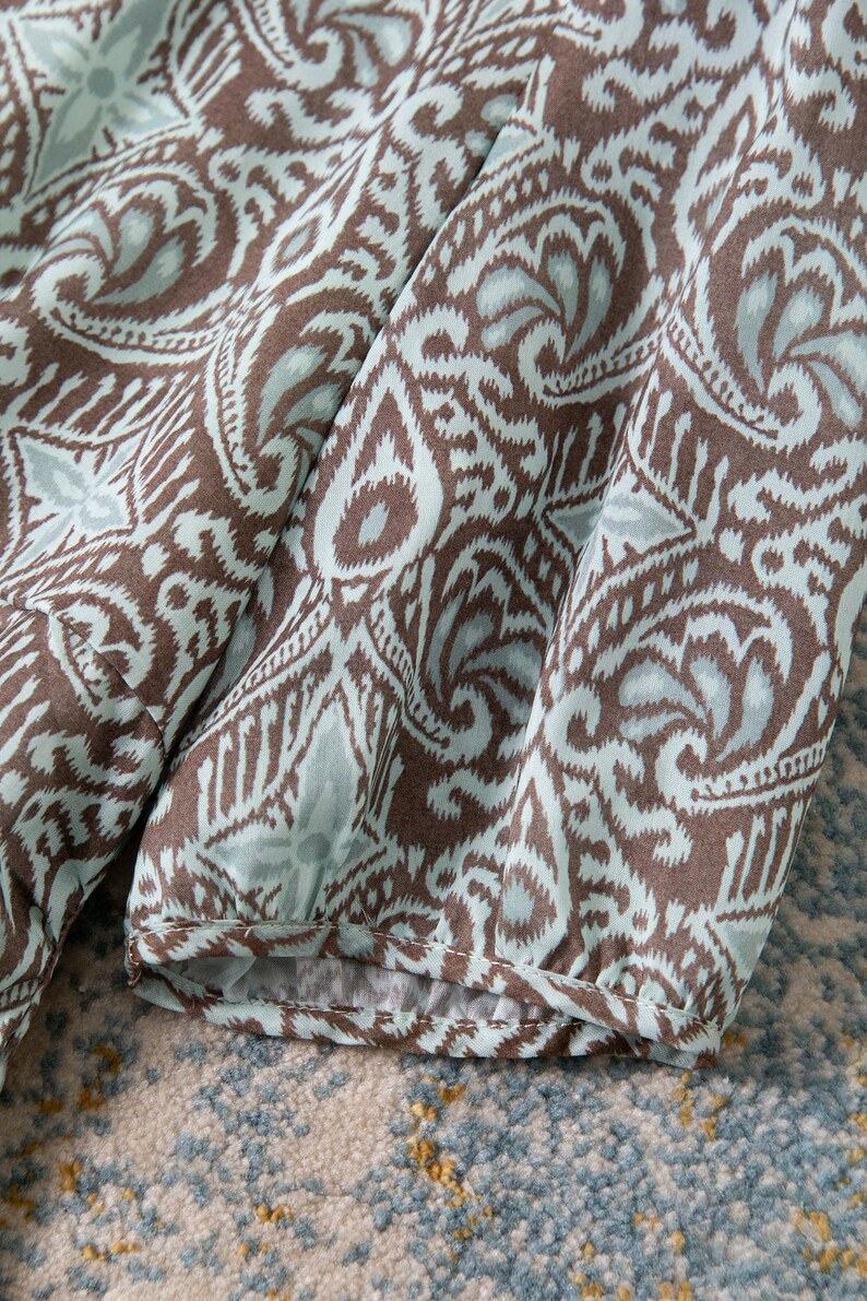 Op de afbeelding: Close-up van stof met een herhalend paisley-patroon in bruin en lichtblauw. De stof is geplooid en toont de mouw van een kledingstuk. Het patroon bevat bloem- en geometrische vormen.