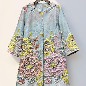 May include: A light blue, knee-length coat with a floral design. The coat features large, embroidered flowers in shades of pink, yellow, and peach. The sleeves are three-quarter length, and the coat hangs on a black hanger.