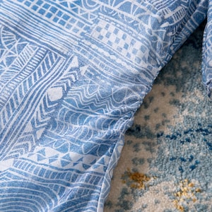 May include: Close-up of a light blue garment with a white tribal pattern. The fabric is gathered along the edge, creating a textured effect. The design features geometric shapes and lines.