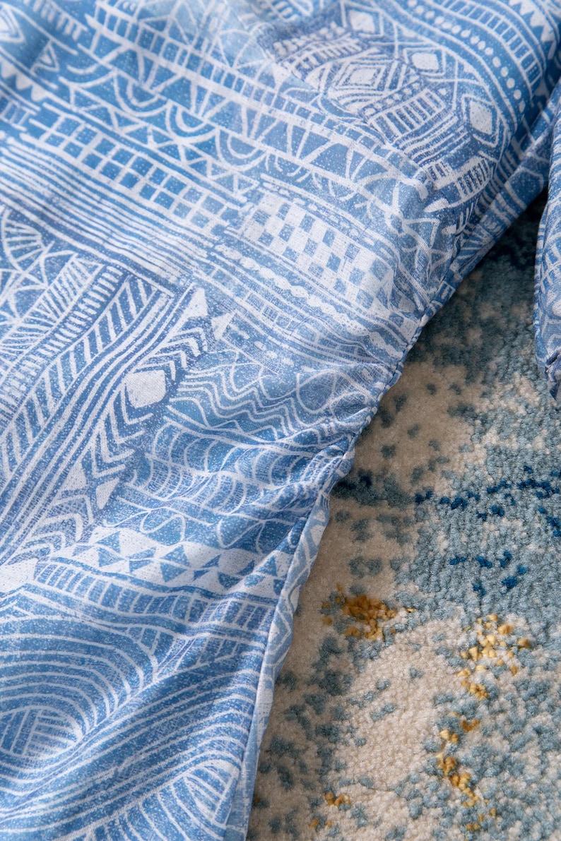May include: Close-up of a light blue garment with a white tribal pattern. The fabric is gathered along the edge, creating a textured effect. The design features geometric shapes and lines.
