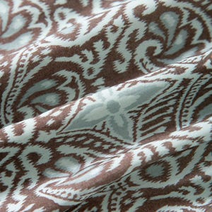 Op de afbeelding: Close-up van een stof met een herhalend paisley-patroon. Het ontwerp heeft bruine en lichtblauwe wervelingen en bloemvormen tegen een lichtblauwe achtergrond. De stof is gevouwen en toont de textuur en de flow van het materiaal.