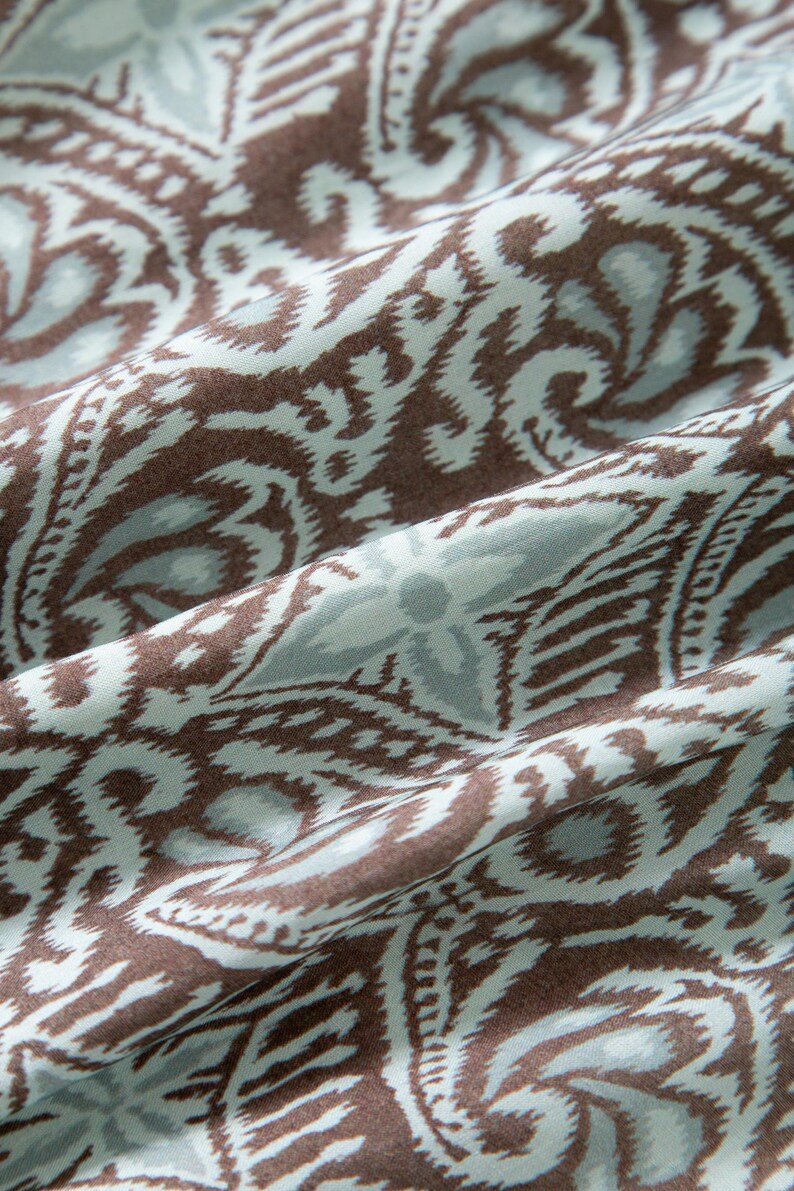 Op de afbeelding: Close-up van een stof met een herhalend paisley-patroon. Het ontwerp heeft bruine en lichtblauwe wervelingen en bloemvormen tegen een lichtblauwe achtergrond. De stof is gevouwen en toont de textuur en de flow van het materiaal.