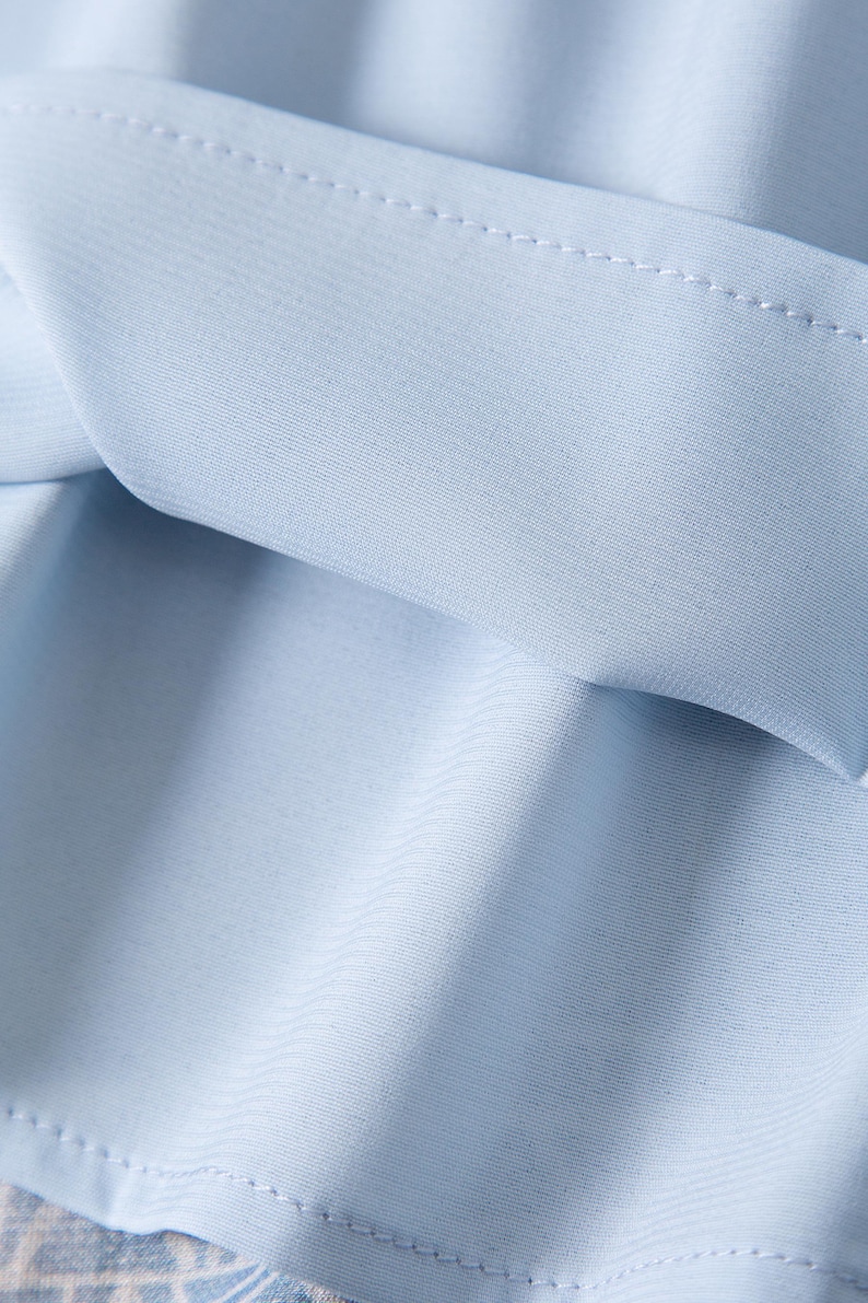 May include: Close-up of light blue fabric with visible stitching along the edges. The fabric appears to be soft and slightly textured, with subtle folds and shadows. The overall impression is of a delicate, flowing material.